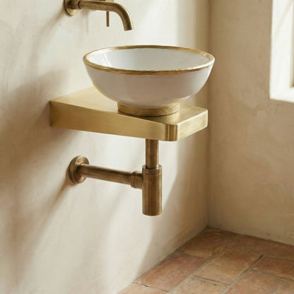 Brushed brass wall-mounted sink bracket supporting white ceramic vessel sink with matching brass faucet and P-trap