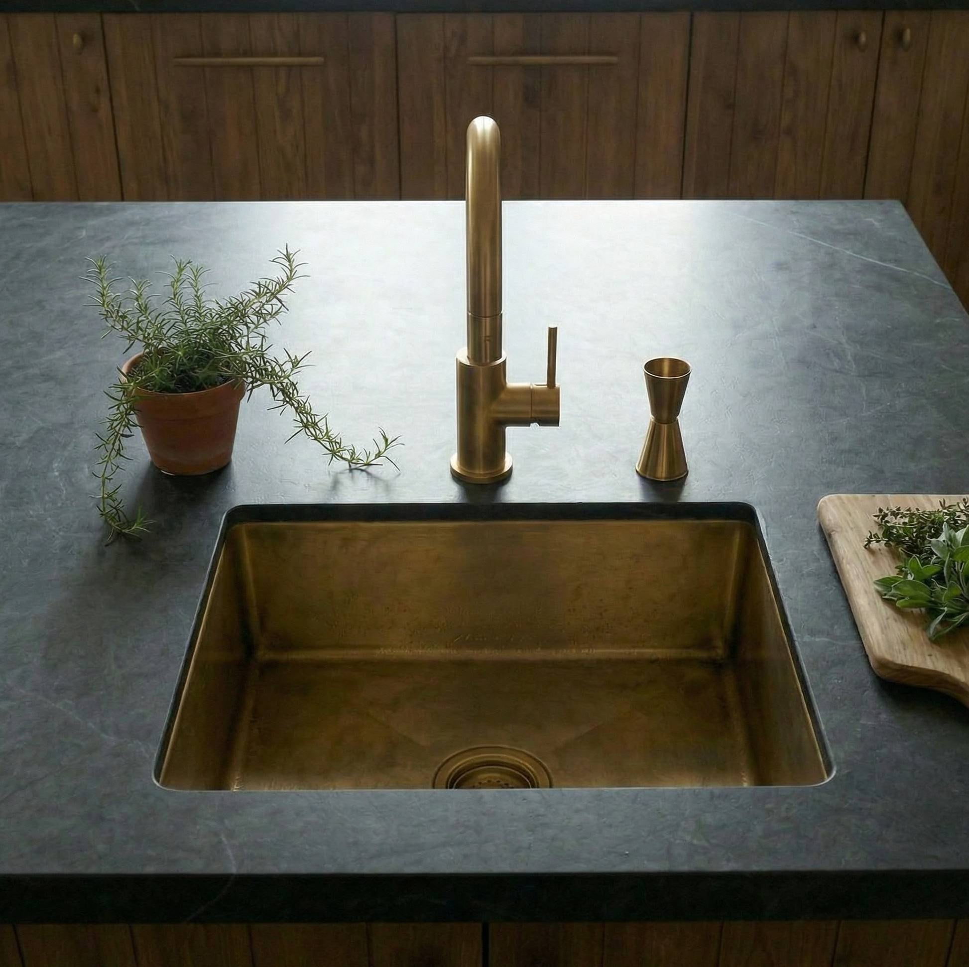 front view brass kitchen sink and faucet on a dark countertop with wooden cabinets in the background.
