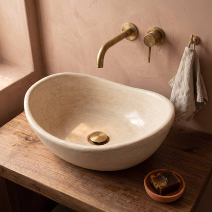 Beige tadelakt vessel sink on a rustic walnut vanity with a brass wall faucet, linen hand towel and black soap in a terracotta dish against a pink plaster wall