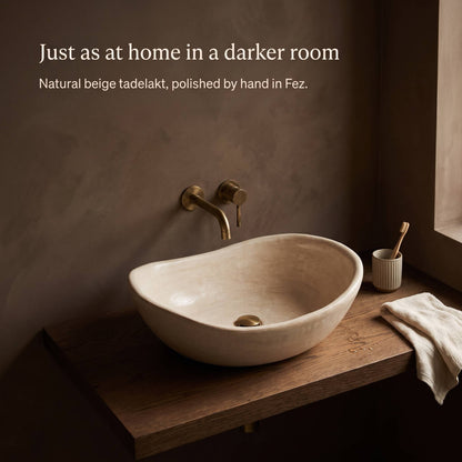 Beige tadelakt vessel sink on a dark oak shelf in a moody brown plaster bathroom, with a brass wall faucet, toothbrush cup and cream towel