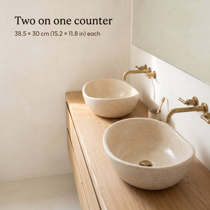 Two small beige tadelakt vessel sinks, 38.5 x 30 cm (15.2 x 11.8 in) each, on a light oak double vanity with brass wall faucets in a cream bathroom