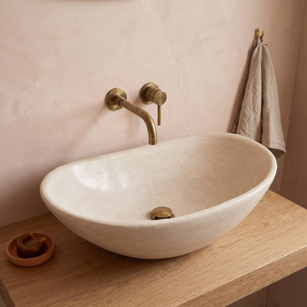 Handmade oval tadelakt vessel sink in natural beige on a light oak shelf, with a wall-mounted brass faucet against a blush plaster wall