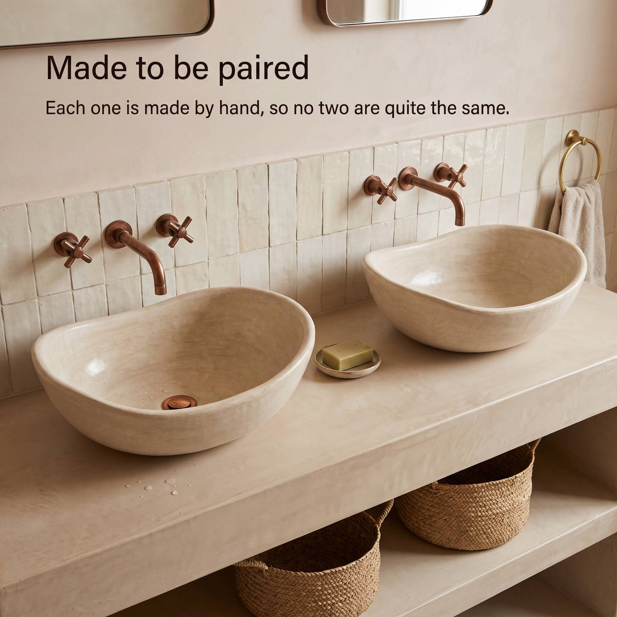 Two handmade beige tadelakt vessel sinks, each slightly different in shape, on a plaster double vanity with copper cross-handle faucets and woven baskets below