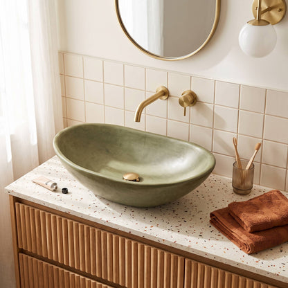 An olive green Tadelakt medium size vessel sink basin, set on a speckled white countertop with a ribbed wood vanity.