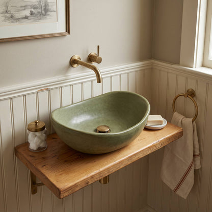 A floating wooden bathroom vanity shelf holding an olive green small Tadelakt vessel sink with a brass faucet, shown in a styled bathroom setting with a towel, soap dish, and wall paneling.