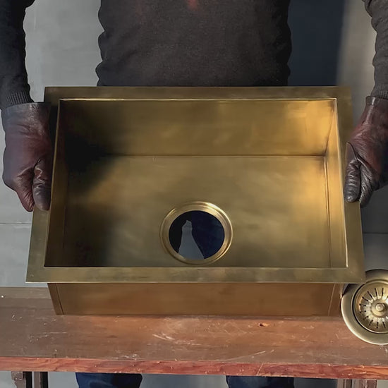 Video showcasing a handmade rectangular aged brass undermount kitchen sink from multiple angles, highlighting the aged bronze patina finish, hand-welded construction, and the included solid brass strainer drain assembly.