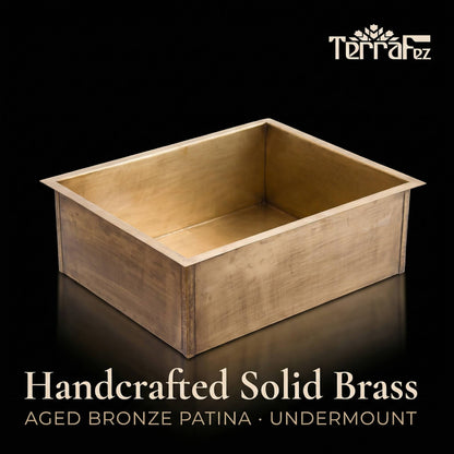 Handcrafted rectangular aged brass undermount kitchen sink by TerraFez, shown from a front three-quarter angle on a black reflective surface, displaying the aged bronze patina finish and single-basin design.