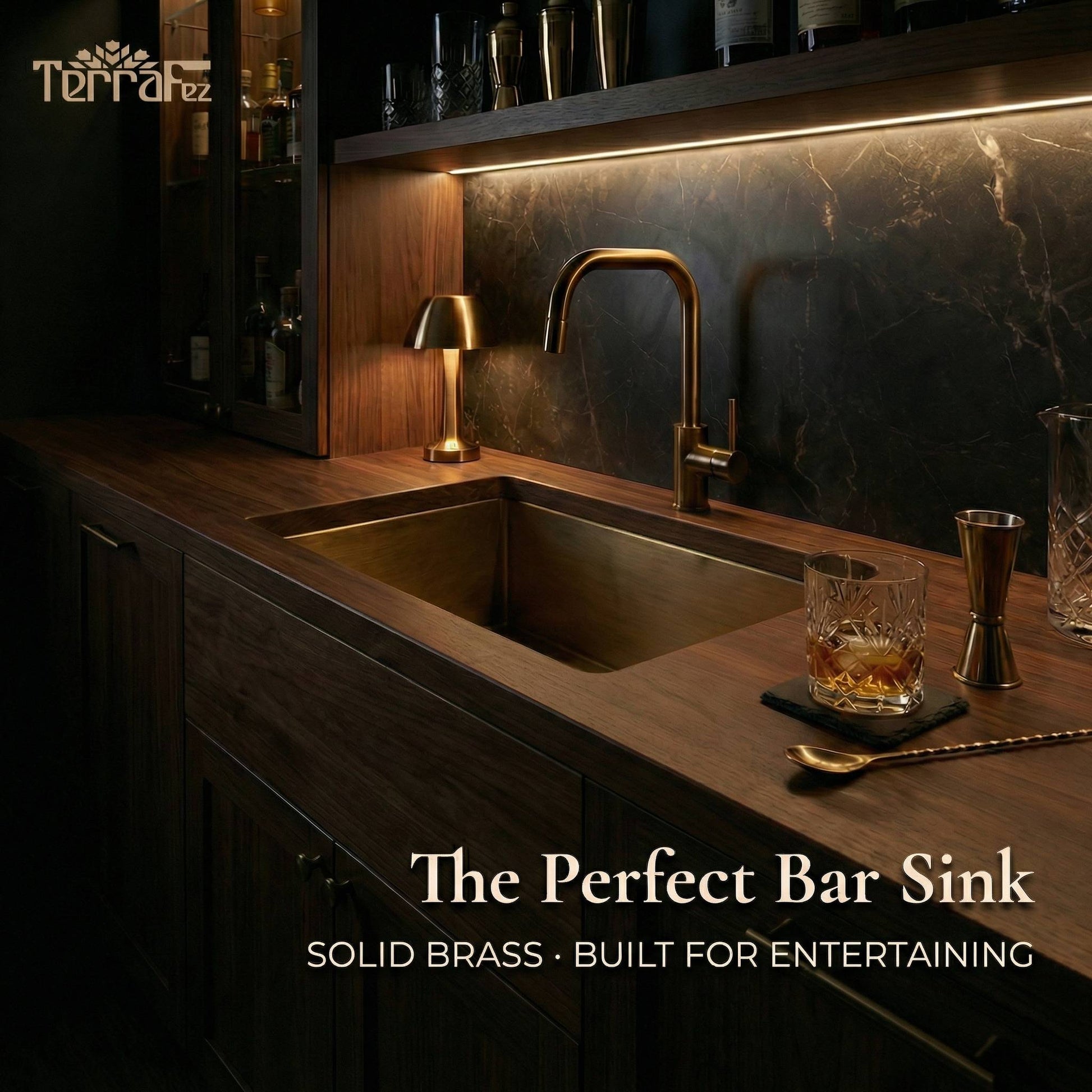 Aged brass undermount sink installed in a dark walnut home bar countertop with a brass faucet, marble backsplash, ambient lighting, and cocktail accessories, showcasing its use as a stylish bar sink for entertaining.
