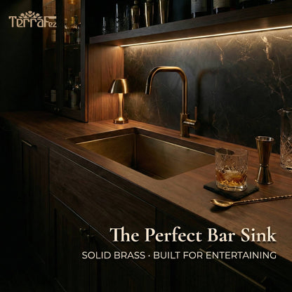 Aged brass undermount sink installed in a dark walnut home bar countertop with a brass faucet, marble backsplash, ambient lighting, and cocktail accessories, showcasing its use as a stylish bar sink for entertaining.