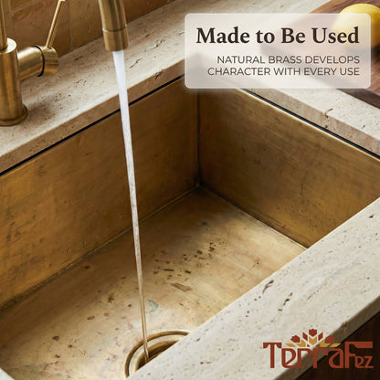 Handmade aged brass undermount kitchen sink installed in a travertine stone countertop with water running from a matching brass faucet, showing the natural patina and character of the brass in everyday use.
