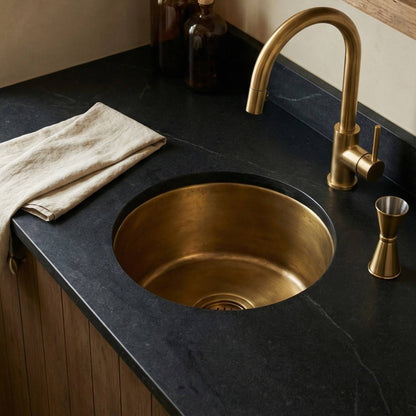 Round undermount aged brass bar sink installed in dark soapstone countertop with matching brass faucet and cocktail jigger at a wet bar