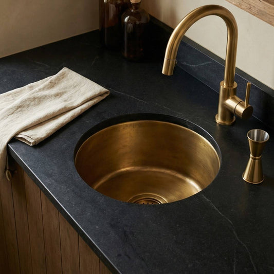 Round undermount aged brass bar sink installed in dark soapstone countertop with matching brass faucet and cocktail jigger at a wet bar
