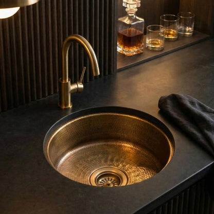 Round undermount aged hammered brass wet bar sink installed in dark countertop with brass faucet, fluted panel backsplash, and whiskey decanter with glasses on shelf