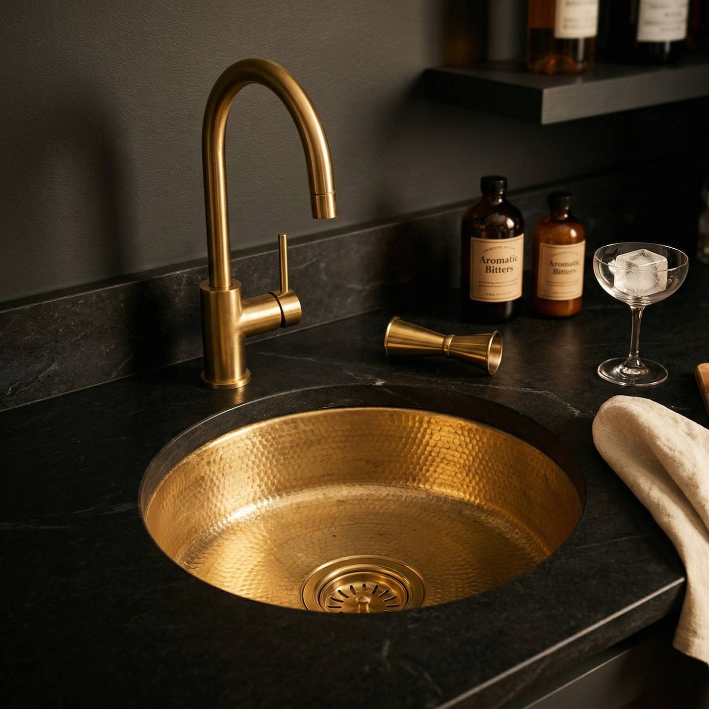 Round undermount hammered brass wet bar sink installed in dark stone countertop with brass faucet, cocktail jigger, bitters bottles, and coupe glass