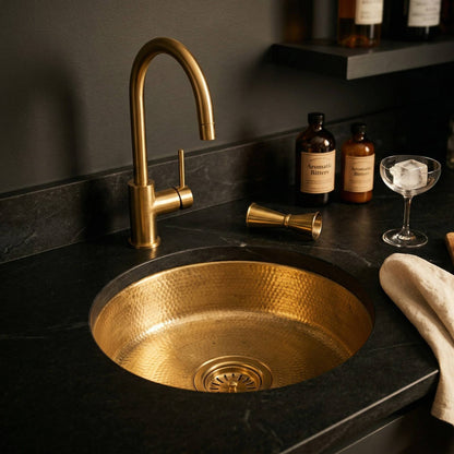 Round undermount hammered brass wet bar sink installed in dark stone countertop with brass faucet, cocktail jigger, bitters bottles, and coupe glass