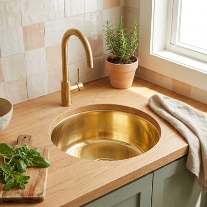 Round undermount polished brass prep sink installed in oak countertop with brass faucet, zellige tile backsplash, and sage green cabinets in a bright kitchen